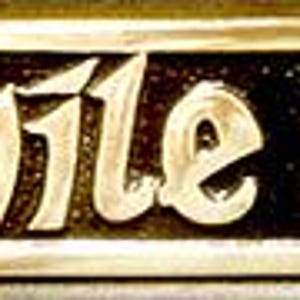 Può includere: Una targa da parete rettangolare in metallo con finitura dorata lucida e sfondo nero incassato. La targa presenta il testo in rilievo "CÉAD MILE FÁILTE" in un carattere stilizzato, con dettagli decorativi a volute alle estremità.