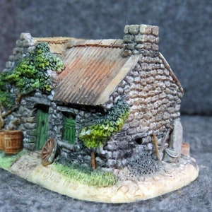 Op de afbeelding: Een klein stenen huisje met een groene deur en een rieten dak. Het huisje is omgeven door groen en heeft een houten vat en een wiel ervoor. De basis van het huisje is beschreven met "Dungloe 1912".