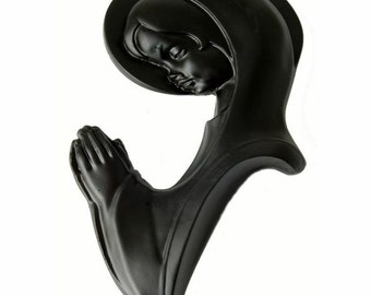 May include: Black painted resin sculpture of a woman in prayer with her hands clasped together. The woman is wearing a robe and has a halo around her head.