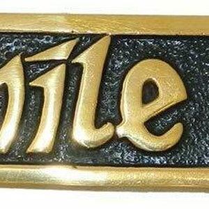 May include: A rectangular sign with the Gaelic phrase "céad míle fáilte" in gold lettering on a black textured background. The sign has a gold border with decorative edges.