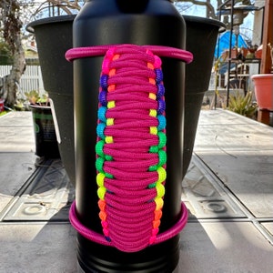 May include: A black water bottle with a pink paracord handle. The handle is woven with rainbow colored paracord.