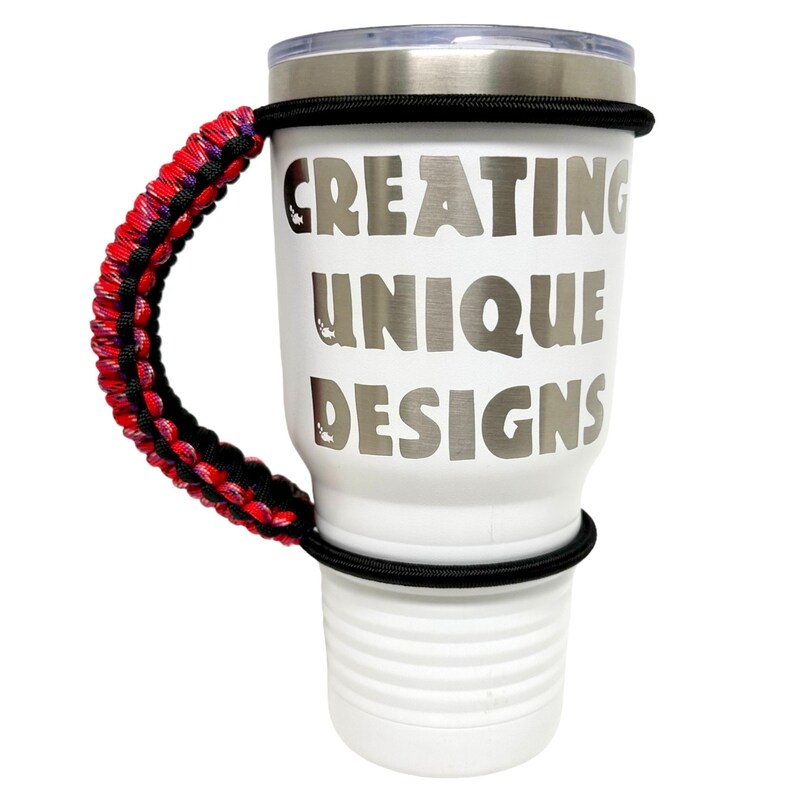 May include: White stainless steel tumbler with a black rubber band and a red, black, and purple paracord handle. The tumbler has the text "CREATING UNIQUE DESIGNS" printed on the side.