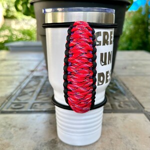 May include: A white tumbler with a black and red paracord handle. The tumbler has the text "CREATIVE UNIQUE DESIGNS" printed on the side.