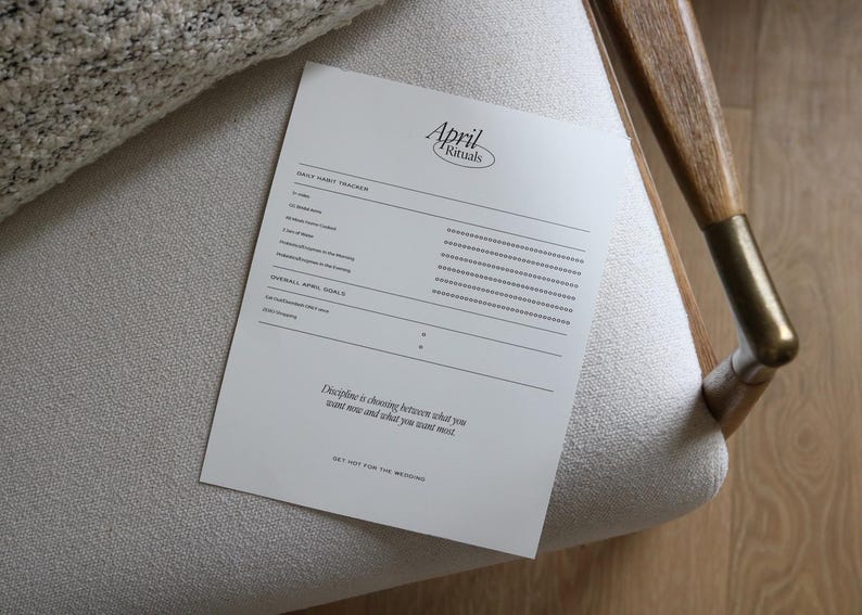 Pu&ograve; includere: Un foglio di carta bianco con il testo "April Rituals" e un tracker delle abitudini quotidiane, appoggiato su una sedia di colore chiaro. La carta include sezioni per il monitoraggio delle abitudini e degli obiettivi generali, con una citazione motivazionale.