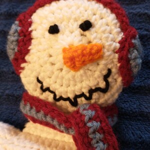 May include: A crocheted snowman with a red and blue scarf and a white crocheted hat. The snowman has black eyes and an orange nose.