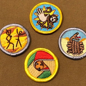Op de afbeelding: Vier kleurrijke geborduurde scout-badges met gele randen. De badges tonen een scène met stokfiguren, een slang, een aap en een hond.