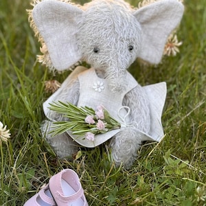 May include: A gray plush elephant teddy bear wearing a white dress with a flower design and holding a bouquet of pink flowers. The teddy bear is sitting on a bed of green grass and has pink shoes.