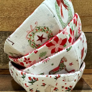 May include: A set of four fabric bowls with a Christmas theme. The bowls are white with red and green accents and feature various holiday designs, including wreaths, poinsettias, and a rocking horse.