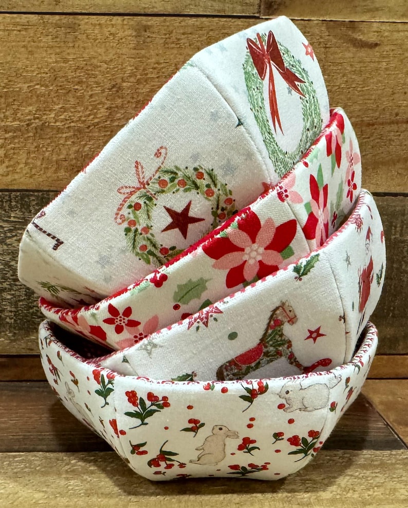 May include: A set of four fabric bowls with a Christmas theme. The bowls are white with red and green accents and feature various holiday designs, including wreaths, poinsettias, and a rocking horse.