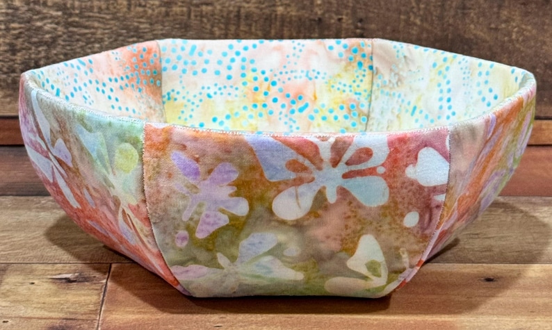 May include: A fabric bowl with a colorful floral pattern in shades of pink, blue, green, and yellow. The bowl is made of fabric and has a unique octagonal shape.