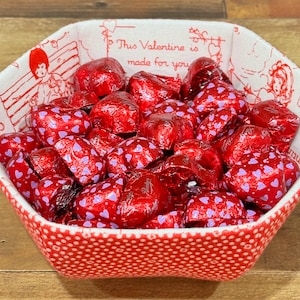 May include: A red and white fabric bowl with a polka dot pattern and a Valentine's Day theme. The bowl is filled with red foil-wrapped heart-shaped chocolates.