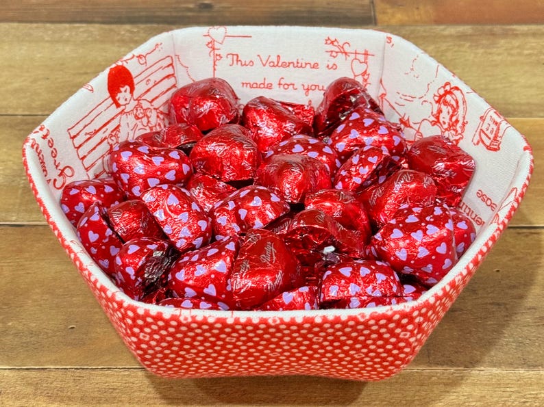 May include: A red and white fabric bowl with a polka dot pattern and a Valentine's Day theme. The bowl is filled with red foil-wrapped heart-shaped chocolates.