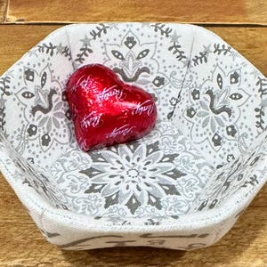 May include: A white fabric bowl with a gray paisley pattern and a red heart-shaped chocolate candy inside.