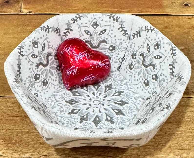 May include: A white fabric bowl with a gray paisley pattern and a red heart-shaped chocolate candy inside.