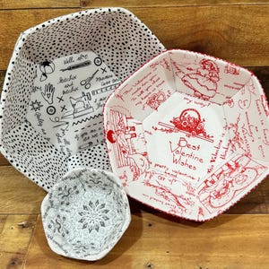 May include: Three fabric hexagon-shaped bowls with white backgrounds and red and black designs. The largest bowl has a red design with the text "Best Valentine Wishes". The medium bowl has a black polka dot design with sewing-themed text. The smallest bowl has a gray and white floral design.