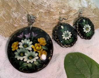 May include: A set of black and gold painted wooden earrings and a pendant featuring a floral design with white and yellow flowers. The earrings and pendant are on a white sandy surface.