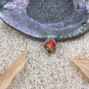 Könnte beinhalten: Ein kleines, ovales, handbemaltes Ornament mit einem Blumenmuster. Das Ornament zeigt rosa, gelbe und grüne Blumen auf weißem Hintergrund. Das Ornament liegt auf einem Bett aus weißem Reis.