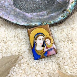 Puede incluir: Una pequeña pintura rectangular enmarcada de la Virgen María y el niño Jesús. La pintura está sobre un fondo blanco con un marco dorado. La Virgen María lleva una túnica azul y una corona dorada. El niño Jesús lleva una túnica rosa y una corona dorada. La pintura está rodeada de arroz blanco.