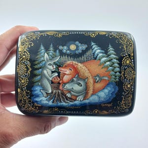 Puede incluir: Una caja lacada en negro con una detallada ilustraci&oacute;n de animales del bosque. Un conejo, un zorro y un lobo se re&uacute;nen alrededor de un toc&oacute;n de &aacute;rbol. La caja est&aacute; decorada con filigranas doradas y el texto "Рукавичка".
