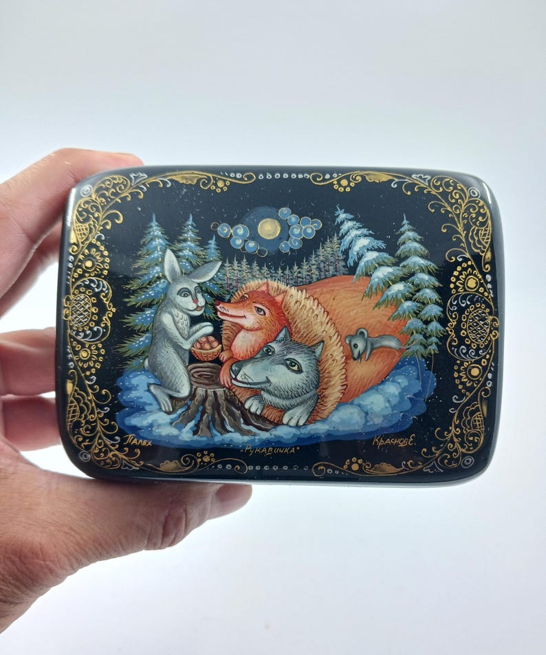 Puede incluir: Una caja lacada en negro con una detallada ilustraci&oacute;n de animales del bosque. Un conejo, un zorro y un lobo se re&uacute;nen alrededor de un toc&oacute;n de &aacute;rbol. La caja est&aacute; decorada con filigranas doradas y el texto "Рукавичка".