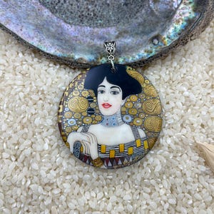 May include: A round pendant necklace featuring a painted portrait of a woman with black hair and a gold background. The pendant is made of a white material with a textured surface.