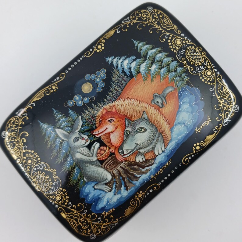 Puede incluir: Una caja lacada en negro con una escena invernal pintada a mano que presenta un zorro, un lobo y un conejo. La caja tiene un borde de filigrana dorada y el texto "Палех" y "Рукавичка".