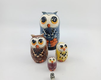 May include: A collection of six owl-shaped wooden nesting dolls. The dolls are painted in shades of blue, brown, yellow, and purple, with large eyes. The largest doll is approximately 15 cm tall.