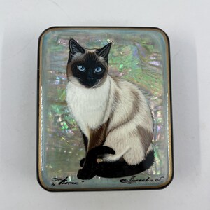 May include: A rectangular, hand-painted trinket box featuring a Siamese cat with striking blue eyes. The cat is depicted in a seated position against a pearlescent, iridescent background. The box has a black border and gold trim.