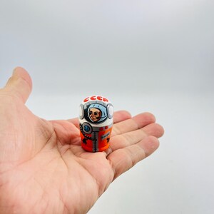 以下が含まれることがあります： 手描きの円筒形の木製人形。宇宙飛行士のデザイン。人形は白を基調とし、赤とオレンジのアクセントがあり、上部には「CCCP」の文字がプリントされています。宇宙飛行士は青いヘルメットと詳細な顔で描かれています。