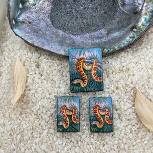 May include: Three rectangular resin cabochons with a painted image of a snake in a jungle setting. The snake is orange and yellow with black markings. The background is green and blue with pink flowers.
