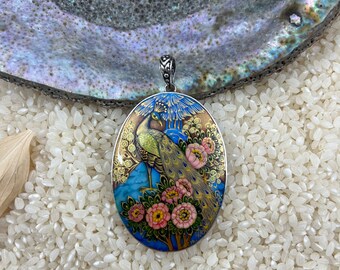 Op de afbeelding: Een ovale hanger met een gedetailleerd schilderij van een pauw met blauwe, groene en gouden veren die op een tak met roze bloemen staat. De hanger is ingelegd in zilver en heeft een zilveren lus.