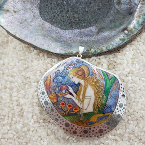 May include: A silver pendant with a painted image of a mermaid holding a pearl. The pendant is surrounded by a silver border with a dotted pattern. The pendant is on a white background.