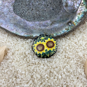 Puede incluir: Un pequeño pin redondo negro y verde con un diseño pintado de dos girasoles amarillos. El pin está sobre un lecho de arroz blanco.
