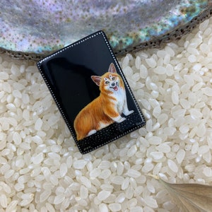 May include: A black enamel rectangular pin with a white border. The pin features a hand-painted image of a smiling corgi dog with orange and white fur.