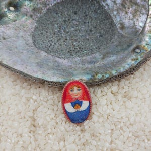 Puede incluir: Una muñeca rusa de madera pintada a mano, o matrioshka, con un diseño rojo y azul. La muñeca sostiene un pequeño objeto naranja en sus manos. La muñeca está sentada en un lecho de arroz blanco.