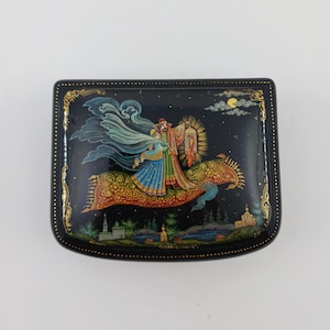 Op de afbeelding: Zwart gelakte doos met een handgeschilderde scène van figuren die op een fantastisch paard door een sterrennacht rijden. De doos heeft een gouden rand en een gedetailleerde illustratie van een stel, een paard en een nachthemel met een maan.