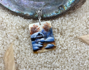 May include: A rectangular pendant with a winter scene painted on it. The scene depicts a small house with snow on the roof and trees in the background. The pendant is made of a white material and has a silver bail.