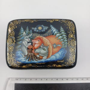 Puede incluir: Caja rectangular lacada en negro con una escena de bosque invernal. Presenta un conejo gris, un zorro rojo y un lobo gris alrededor de un toc&oacute;n. La caja est&aacute; decorada con filigranas doradas y el texto "Рукавичка".
