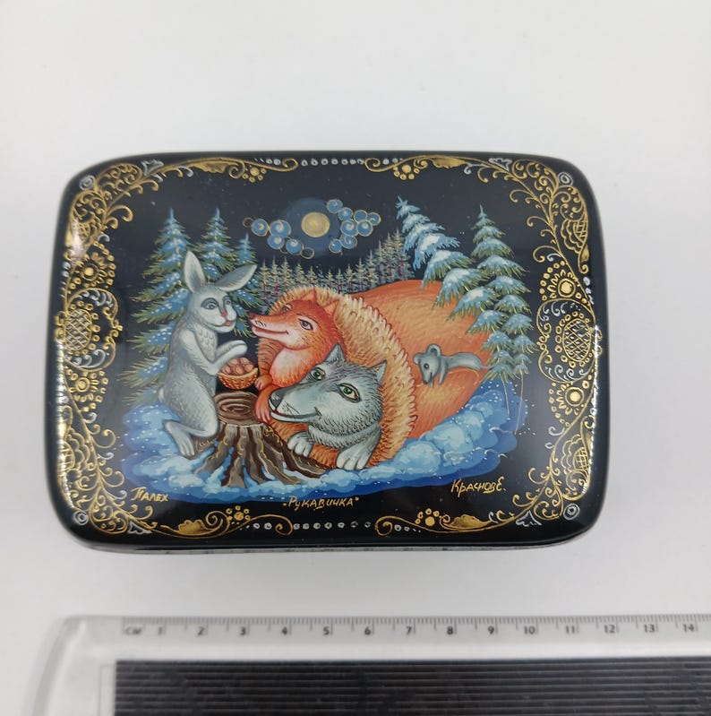 Puede incluir: Caja rectangular lacada en negro con una escena de bosque invernal. Presenta un conejo gris, un zorro rojo y un lobo gris alrededor de un toc&oacute;n. La caja est&aacute; decorada con filigranas doradas y el texto "Рукавичка".