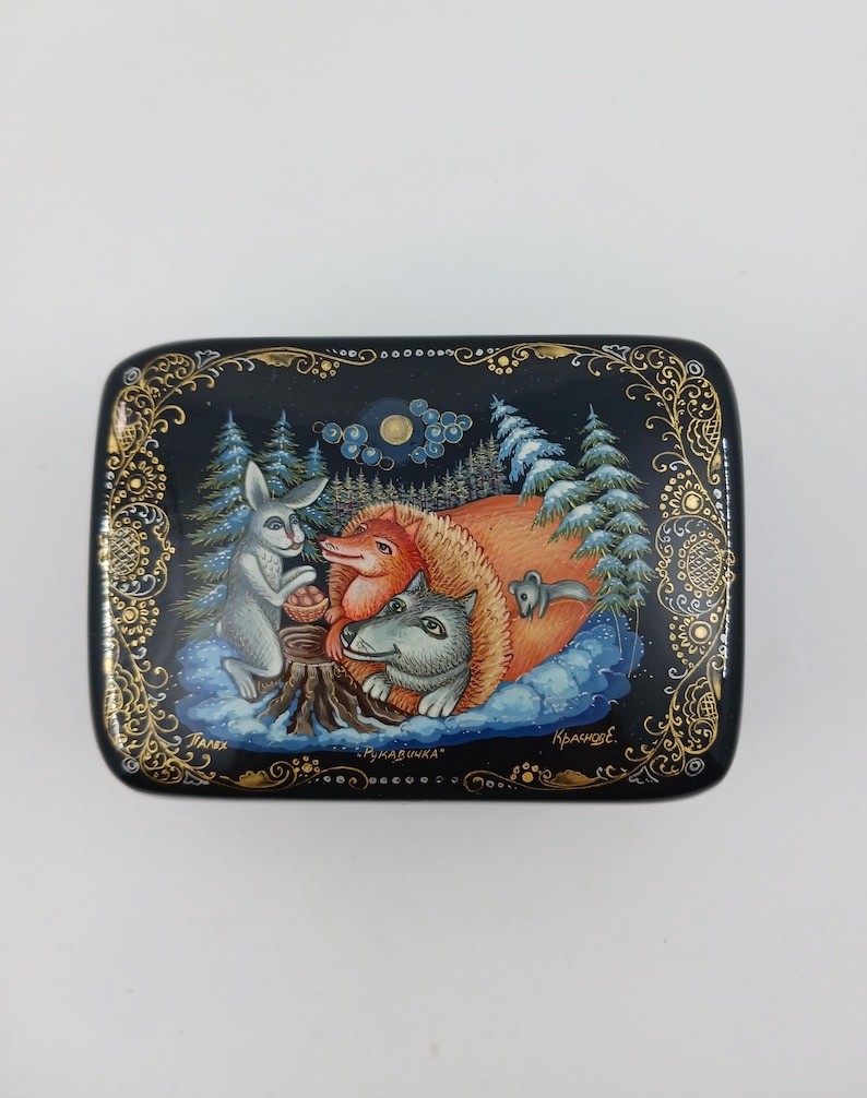Puede incluir: Caja rectangular negra con una pintura detallada de una escena de bosque invernal. Se representan un conejo gris, un zorro rojo y un lobo gris. La caja tiene bordes dorados y texto en ruso.