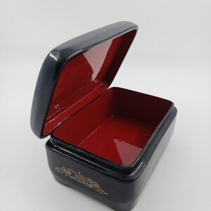 Puede incluir: Una peque&ntilde;a caja negra rectangular con tapa abisagrada, abierta para revelar un interior rojo. La caja tiene detalles decorativos dorados en la parte delantera y los lados. La caja probablemente se utiliza para guardar objetos peque&ntilde;os.