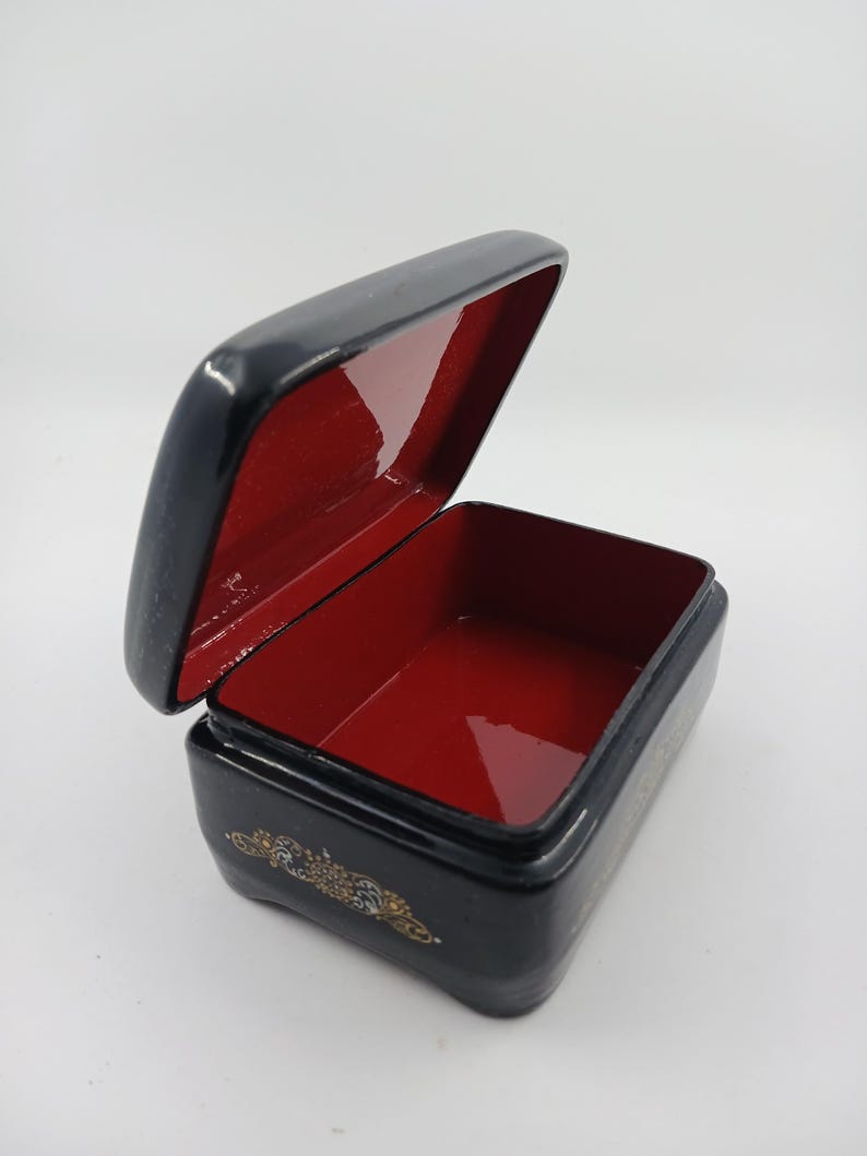 Puede incluir: Una peque&ntilde;a caja negra rectangular con tapa abisagrada, abierta para revelar un interior rojo. La caja tiene detalles decorativos dorados en la parte delantera y los lados. La caja probablemente se utiliza para guardar objetos peque&ntilde;os.