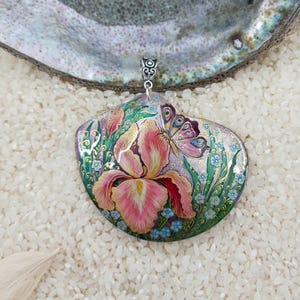 May include: A hand-painted pendant in the shape of a seashell, featuring a pink iris and a butterfly. The pendant has a silver bail and is set against a background of white rice.