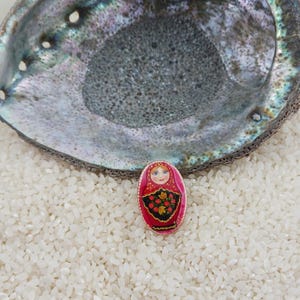 Puede incluir: Un pequeño broche de muñeca rusa, de forma ovalada, con un pañuelo rojo y un vestido negro adornado con motivos florales rojos. El broche descansa sobre arroz blanco, con una gran concha iridiscente encima.