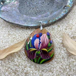 May include: A hand-painted pendant featuring a purple and pink iris flower with green leaves and a red ladybug. The pendant is made of a light brown stone and has a silver bail.