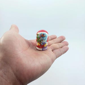 Könnte beinhalten: Eine kleine, handbemalte Holz-Matrjoschka-Puppe mit einem Weihnachtsmann-Design. Die Puppe hat einen roten Hut und Mantel, mit einem blauäugigen Weihnachtsmann, der einen grünen Weihnachtsbaum hält. Die Puppe wird in der Hand einer Person vor weißem Hintergrund gehalten.
