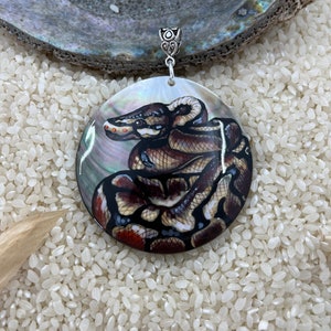 May include: A round, iridescent, mother-of-pearl pendant with a painted image of a brown and black snake coiled up. The pendant has a silver bail.