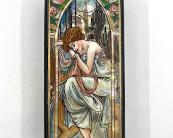 May include: Rectangular black box with a decorative Art Nouveau design. The box features a painting of a woman with flowing auburn hair, wearing a flowing gown, and resting her head on her hand. The background includes a stained-glass window design with pink flowers.