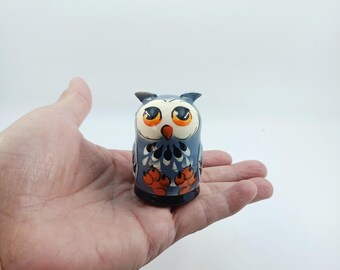 May include: A small, hand-painted wooden owl figurine featuring a blue body, large orange eyes, and orange feet. The owl has a cream-coloured face with dark blue feather details on the chest and a black base, presented as a single decorative object.