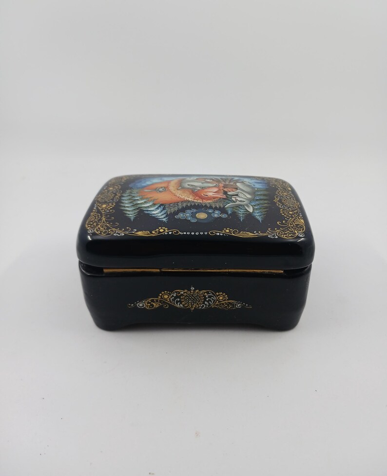 Puede incluir: Una caja negra lacada con tapa con bisagras. La parte superior presenta una pintura detallada de una escena de bosque con un zorro y otros animales. Los bordes de filigrana dorada bordean la parte superior e inferior de la caja.